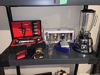 Wide shot showing the assembled lot on a plastic shelving unit including blender, boxed glassware, flask, bartending kit, and additional glasses.