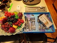 Photo of ceramic bowl filled with realistic faux fruits including red grapes, pomegranate, green apple, purple grapes, lemon as well as boxed Lagostina cutlery set containing forks, spoons, and knives; butter dish also visible.