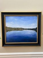 Framed oil painting showing overall view of the lake landscape with blue sky and dark green treeline reflection on water.