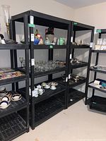Photo showing two black plastic shelving units filled with various items including glassware, ceramics, and silverware, indicating shelving units' condition and size.