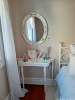 White oval framed wall mirror hung on beige wall above white side table with detailed legs, decorated with books and figurines (not included).