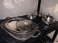 Photo showing large rectangular and oval silver plate trays, teapot, covered butter dish, and soup bowl with ladle on a black metal shelf.