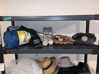 Shelf with Broadstone sleeping bag in blue carrying bag, RBZ driver golf club, Legendary Motorcar Company black cap, Definity resistance cord package, two baseball gloves (tan and black) with baseballs on them and additional baseballs on the shelf
