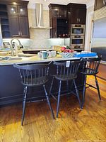 Three black wooden stools with backs placed at a kitchen counter with dark cabinetry and hardwood floor.