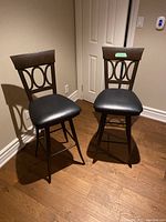 Front view of two stools showing black vinyl padded seats, wood backrests with circular cut-outs, and dark metal frames.