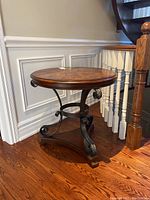 Full view of round wood table with burl wood inlay top and metal scrolling base, placed on wood floor next to white staircase