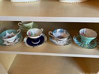 Photo showing 5 teacups each sitting on their matching saucer, lined up on a shelf. Various colors and patterns with gold accents.