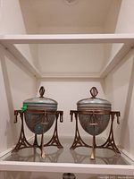 Front view of two light blue porcelain urns with bronze stands and lids, displayed side by side on glass shelf