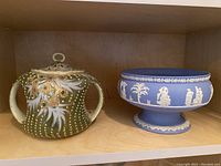 Shelf showing two items: ornate hand-painted jar with loop lid handle and two handles on sides, mainly green with gold and white decoration; next to it, a blue Wedgwood footed bowl with classical white relief decoration.