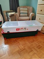 Three rectangular plastic underbed storage boxes stacked on parquet floor, near armchair and chest of drawers. Each box has a white lid and wheels, colored bases vary.