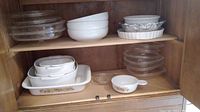 Overview photo showing multiple stacked Corning Ware pieces on wooden shelf including white and floral patterned dishes, clear glass bowls, and lids.