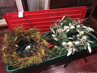 Photo showing two wreaths inside a storage container, one decorated with pinecones and berries, the other with silver and gold ornaments.