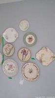 Photo showing nine assorted decorative collector plates with floral designs and different shapes, including round and square plates, some with gold trim and ornate edges.