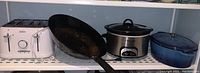 Full shelf view including Sunbeam 4-slice toaster, black wok, Crockpot slow cooker, and blue cast iron casserole baking dish.