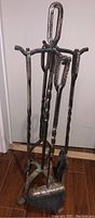 Full view of metal fireplace tool set on stand showing four tools including brush, shovel, poker, and tongs with rustic metal finish and some rust marks.