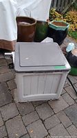 Suncast plastic garden hose box outside on patio with green garden pots in background, towel covered couch partially visible.