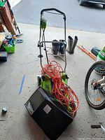 Greenworks lawnmower with handlebar, orange extension cord wrapped around it, wheels visible, and rear grass catcher bag with brand name.