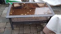 Photo of rectangular faux wicker patio coffee table showing the wet glass top surface with leaves and debris on it, placed on a paved outdoor area surrounded by outdoor furniture covered with protective covers.