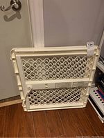 Off-white plastic pet gate in folded position showing lattice panel and label with instructions.