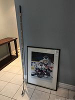 Full view photo showing framed hockey goalie print and hockey stick leaning against a wall, set on tile floor.