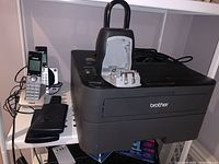 Brother black multifunction printer with Master Lock combination padlock placed on top, lock is open. Also visible are two VTech cordless handsets and charging base.