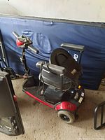 Photo showing the full scooter with seat and armrests, red and black frame, in storage area.