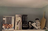 Photo of shelf containing all six appliances and wooden pizza board showing Cuisinart bread maker box, pressure cooker, blender, mixer, kettle and wooden board