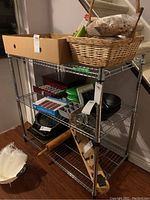 Steel metal rack with 3 wire shelves filled with various household items including baskets, cardboard box, trays, bowls, and miscellaneous kitchen items.