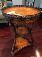 Full view of a round wood side table with metal edge gallery and three wooden shelves on curved legs, showing wood grain and minor wear.