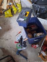 Overall view of an open blue toolbox with various tools and containers inside, showing a partially visible black handled tool and red handled screwdriver on floor.