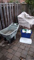 Wide view showing resin wheelbarrow, covered furniture, and box with garden tools arranged on patio with stone tiles and wooden fence