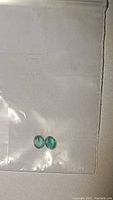 Pair of two small oval emerald gemstones on white background inside plastic packaging.