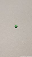 Close-up photo of a small oval cabochon cut tsavorite garnet gem with rich green color against a plain background.