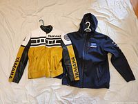 Two Yamaha men's jackets side by side: one black hooded FX racing jacket and one yellow, white, black leather racing jacket with Yamaha branding.