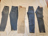Five pairs of men's jeans laid flat on bed showing variety of brands, sizes, colors; good and worn condition