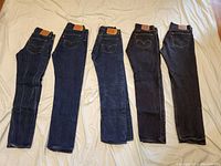 Five pairs of Levi's 501 jeans laid flat, showing front and back with tag patches visible. Two pairs appear black or very dark indigo, three pairs dark blue indigo.