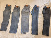 Photo shows five pairs of men's denim jeans laid out flat side-by-side, showing front and side views. Each pair has the characteristic Levi's or Lucky Brand leather patch on the back waistband. Denim washes vary from light to medium blue. Jeans show visible signs of wear including fading and creasing.