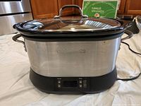 Oval-shaped Calphalon digital slow cooker with stainless steel body and glass lid on a kitchen surface, showing front digital control panel and brand name.