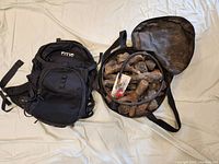 Photo showing side-by-side items on beige fabric: RTIC black hydration pack and Camp Chef fire pit kit with synthetic logs and hose in open carrying case.