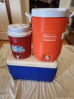 Photo of three coolers placed on the floor including two Rubbermaid water coolers (one large orange and one smaller red) and a large blue Igloo cooler at the bottom.