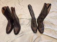 Photo showing two pairs of men's cowboy boots side by side on a light fabric surface. Left pair: Tecovas dark brown ostrich skin boots, unworn. Right pair: dark brown stitched design boots, unknown brand, worn.
