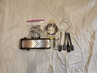 Photo showing the Calphalon Precision Mandoline Slicer with blade accessories in plastic bag, 4 silicone spatulas in gray and black, small yellow colander, Pyrex measuring cup, and glass jar butter churn/fermenter with metal lid, all laid out on a white cloth.