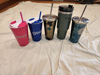 Photo of 5 drink containers displayed on cloth backdrop showing two plastic 'geek' tumblers, two metal Starbucks tumblers, and one Stanley insulated tumbler.