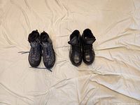 Photo showing two pairs of men's boots placed side by side on a light fabric surface. Left pair is blue and black hiking boots, right pair is black leather leather boots.