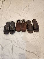 Photo showing three pairs of men's slides and slippers arranged side by side on a white fabric background. Includes black Footjoy slides, brown Hi-Tec slippers, and brown Golukai slippers.