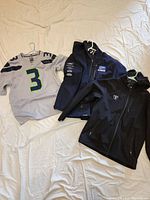 Three items laid flat, grey Nike NFL Wilson jersey with number '3', black hooded Yamaha racing jacket with logos on sleeve, and black hooded RacerX jacket