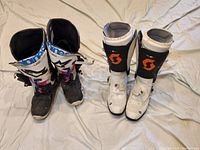 Photo showing two pairs of motocross motorcycle boots on a light surface. Left pair is Alpinestars LXIII black with blue, pink and white accents. Right pair is Scott 550 white with black and orange logo.