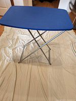 Small folding utility table standing on a beige cloth, blue rectangular top with silver metal frame and folding legs visible.