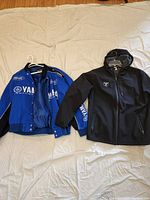 Two men's jackets laid flat on a bed: blue Yamaha letterman style jacket on the left and black RacerX hooded jacket on the right.