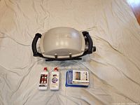 Photo of Weber portable propane grill on white sheet with two bottles of lighter fluid and plastic container of fire starters in front